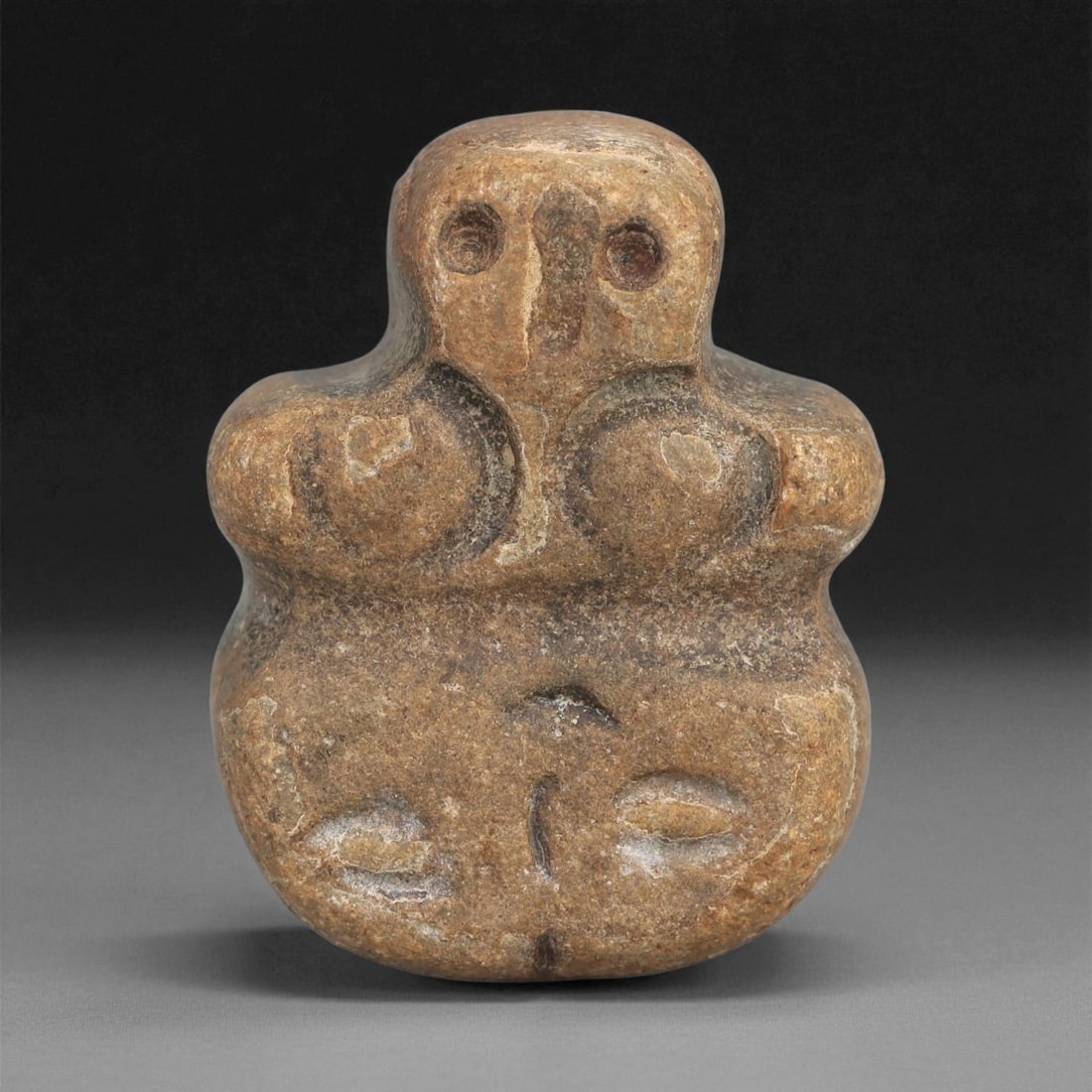 A Near Eastern Stone Anthropomorphic Figurine, 4th-3rd Millennium BCE, 16LD: Carved from a brown stone with smooth surface wear, the figurine is modeled with a rounded head, drilled circular eyes, and a narrow vertical nasal ridge. The upper body is broad and features two pron