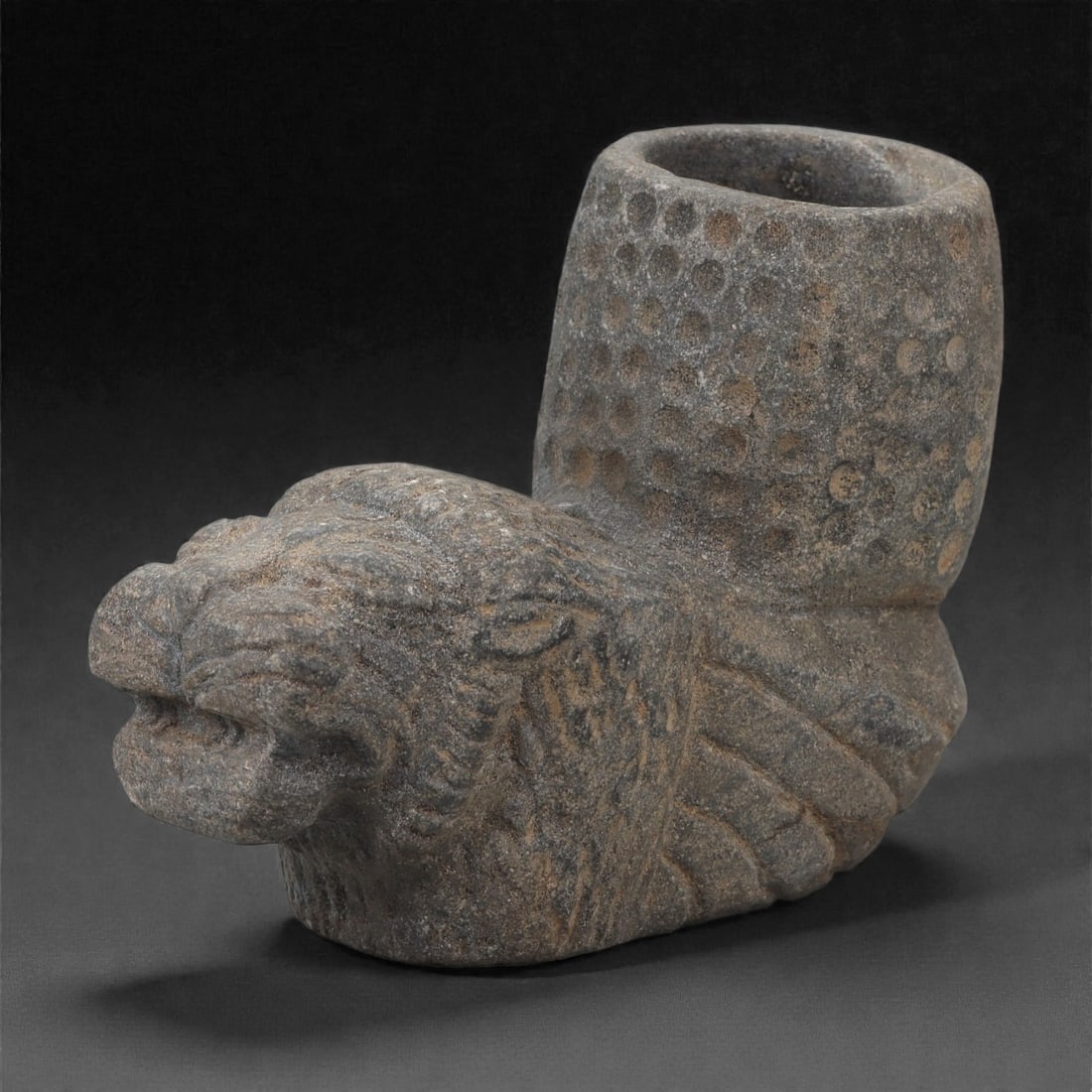 An Anatolian Grey Stone Lion-Head Vessel, Iron Age, 8th-7th Century BCE, 8LM: Carved from a dark grey stone with a granular texture, the vessel is formed with a cylindrical cup rising behind a projecting lion head. The head is modeled with a broad snout, defined nostrils, and i