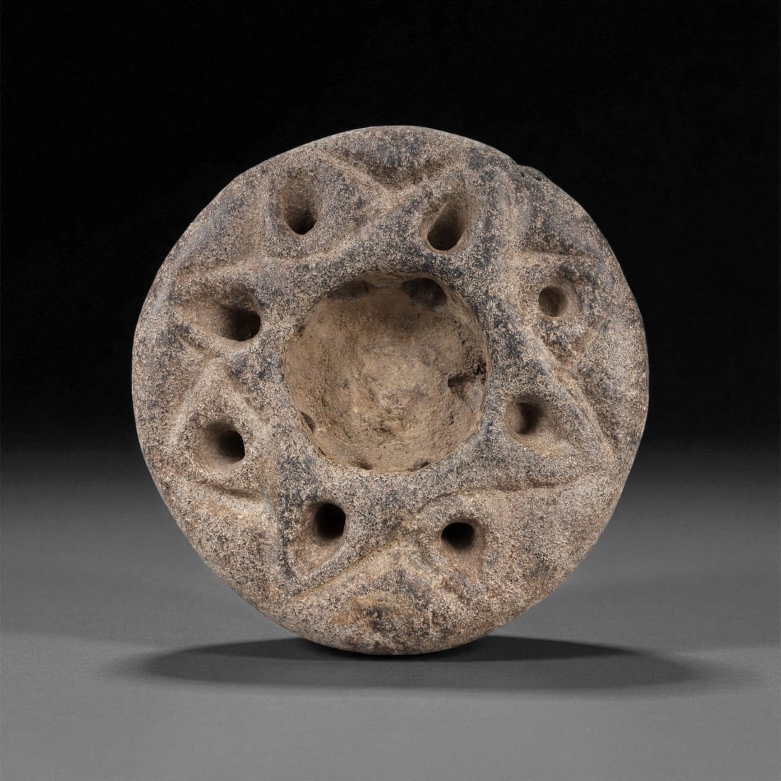 An Eastern Iranian Stone Votive Vessel, Jiroft Culture, 3rd Millennium BCE, 8LE: Carved from a grey-brown stone with surface encrustations, the small circular vessel has a deep central recess surrounded by a broad rim. The rim is decorated with a continuous band of incised geometr