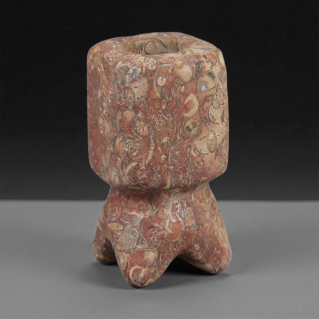 A Near Eastern Stone Kohl Vessel, 2nd-1st Millennium BCE, 25LE: Carved from a red and beige fossiliferous stone, the vessel has a cylindrical upper chamber with a narrow central aperture and a flaring lower section supported on four short lobed feet. The exterior