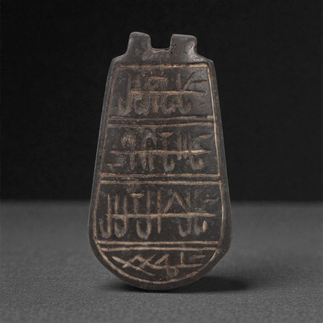 A Mesopotamian Steatite Plaque Amulet, Early Dynastic to Akkadian, c. 2600-2200 BCE, 89LN: Carved from dark grey steatite, the amulet is shaped as a tapering plaque with a rounded lower end and two upright lugs at the top forming the suspension feature. The front surface is divided into fou