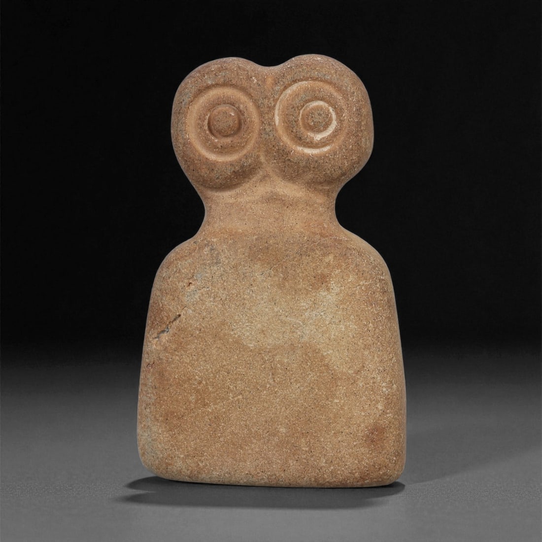 A Near Eastern Stone Eye Idol, 4th-3rd Millennium BCE, 96ZB: Carved from a light brown stone with fine surface inclusions, the figure is shaped with a rounded upper section bearing two recessed circular eyes defined by concentric grooves. The shoulders slope ge