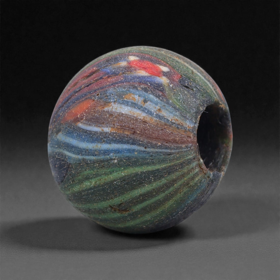 Ancient Roman Wound Glass Bead - Multicolored Marbled Sphere, 23J: Shaped through the winding of molten glass around a mandrel, this small bead displays a marbled surface composed of flowing bands in green, blue, red, white, and brown. The swirling coloration reflect
