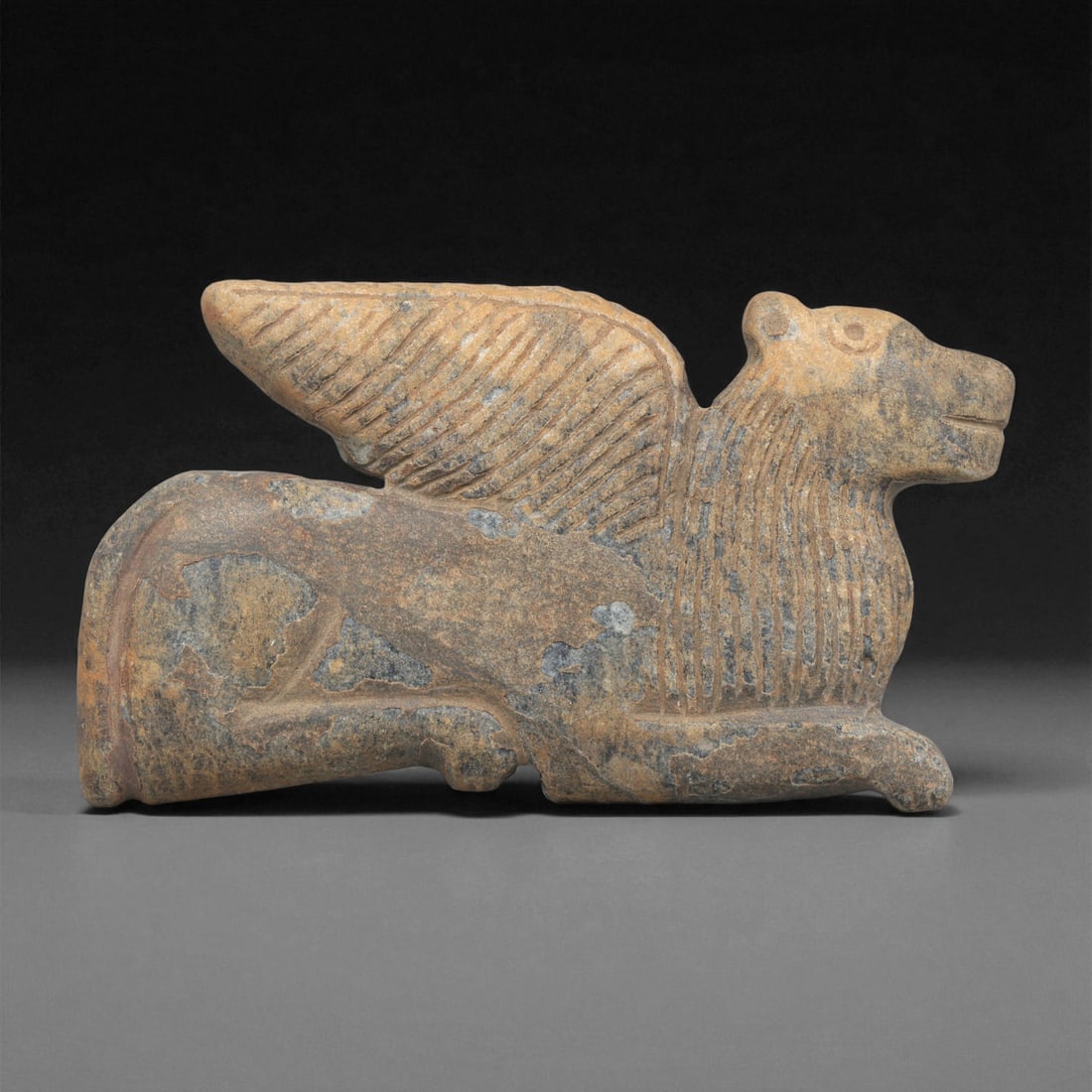 An Indo-Parthian Stone Winged Creature Plaque, 1st-3rd Century CE, 30ZA: Carved from a light brown stone with darker surface accretions, the plaque depicts a recumbent winged creature in profile. The head is rendered with a rounded muzzle and small ears, while the chest an