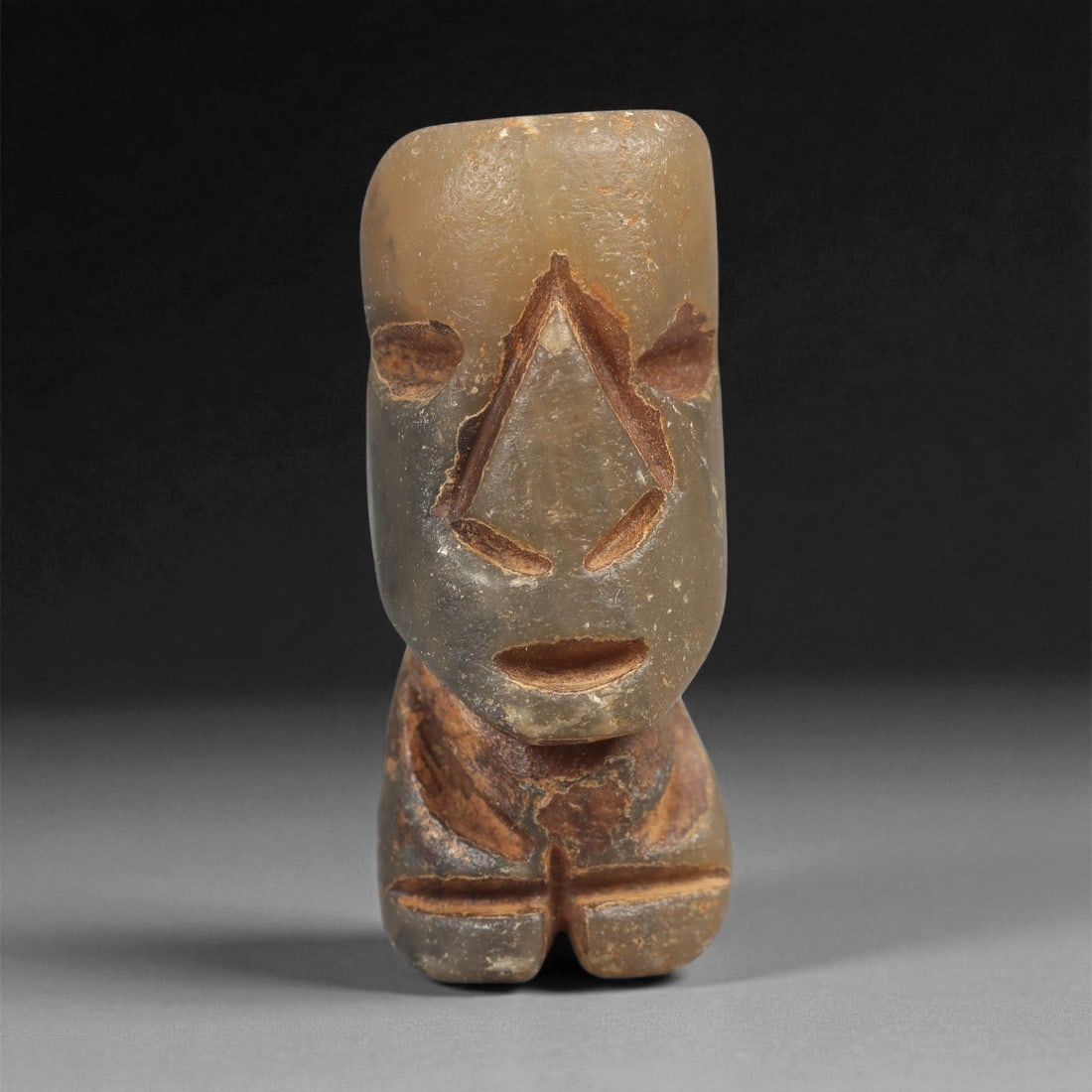 A Taino Carved Stone Idol, Late Ceramic Age, circa 1200-1500 CE, 33LD: Carved from a translucent brown stone with reddish surface deposits, the figure is shown standing with short legs and a compact body. The head is elongated and smoothly rounded above a sharply incised