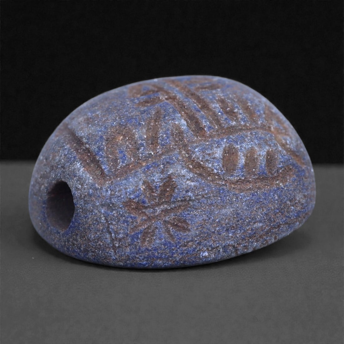 A Parthian Lapis Lazuli Engraved Bead, Circa 1st Century BCE-2nd Century CE, 1N: Carved from lapis lazuli with mottled deep blue coloration and lighter mineral inclusions, the bead is ovoid in form and pierced longitudinally for suspension. The exterior surface is engraved with a 