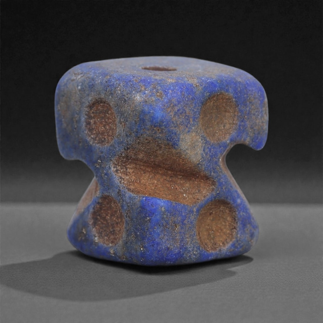 A Phoenician Lapis Lazuli Bead, Circa 7th-5th Century BCE, 100L: Carved from lapis lazuli exhibiting a mottled deep blue surface with lighter mineral inclusions, the bead is compact in form with a thickened central body and recessed lateral contours. The surface is