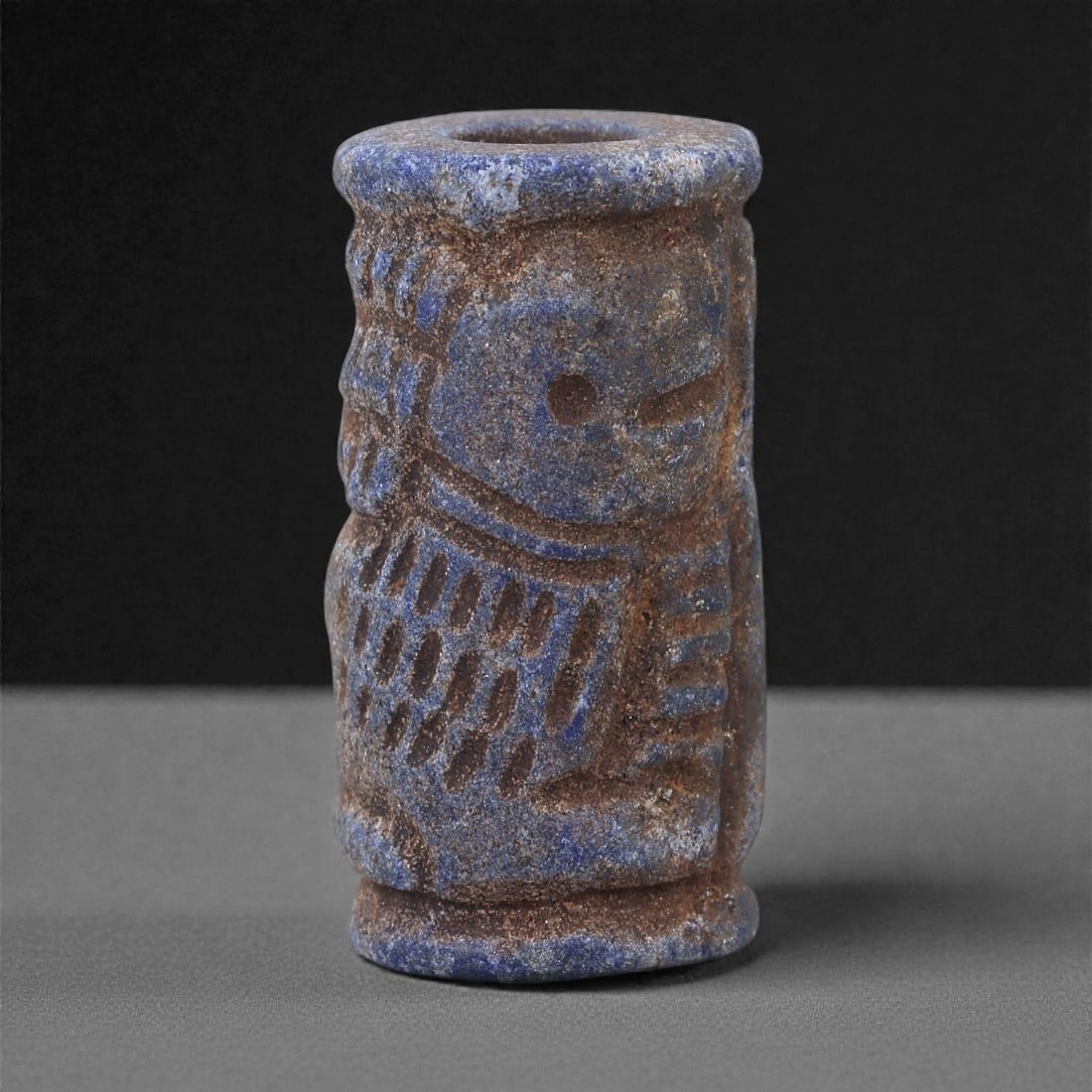 A Near Eastern Lapis Lazuli Cylindrical Relief Amulet, 3rd Millennium BCE, 2.9cm, 92L: Carved from lapis lazuli with mottled deep blue coloration interspersed with lighter calcite and brown mineral inclusions, the object is cylindrical in form with flat upper and lower faces. A longitud