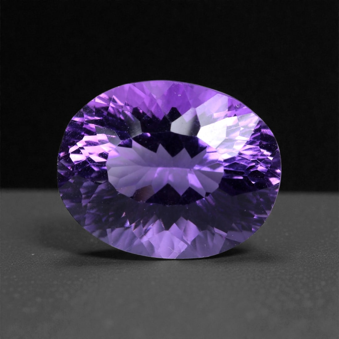 10.57 Carats Violet Amethyst Oval - Natural Brazilian Gem, Concave Cut, 83XB: This radiant amethyst exudes a vivid violet hue with a striking internal brilliance enhanced by a modern concave cut. Measuring 15.9 x 12.3 x 9.6 mm, the stone's expertly crafted facets create a dynam