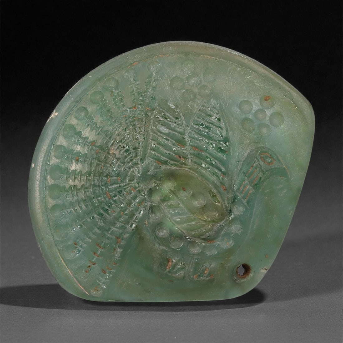 A Sasanian Green Glass Plaque with Peacock, 3rd-7th Century CE, 32LP: Cast from translucent pale green glass, the plaque is shaped as an asymmetrical rounded form and decorated in low relief with a peacock in profile. The bird is shown with an arched neck, a rounded bod