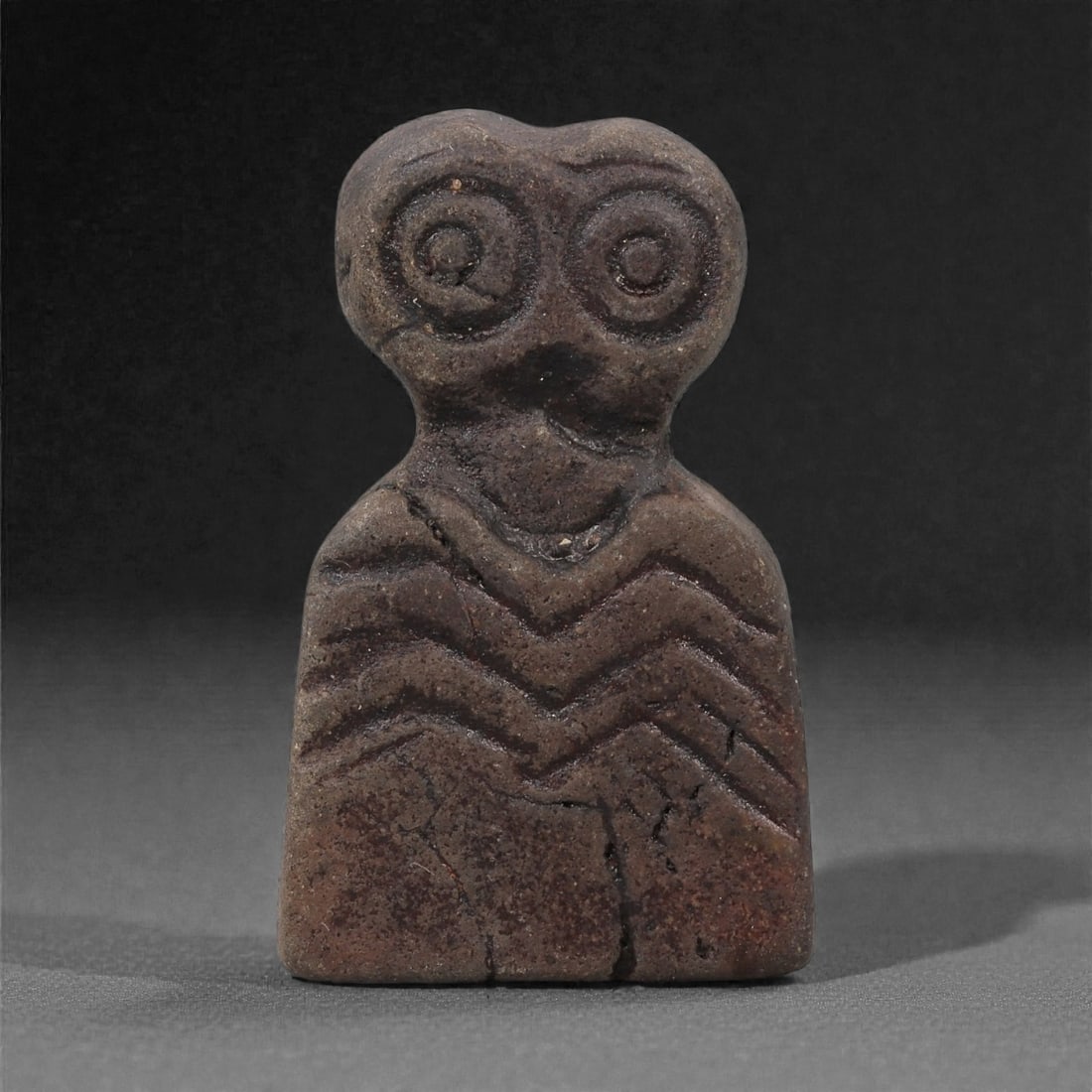 A Near Eastern Stone Eye Idol, 4th-3rd Millennium BCE, 53LH: Carved from a dark brown stone with a finely granular surface, the figure is shaped with a squared lower body and a narrow neck supporting a twin-lobed head. The eyes are rendered as large concentric