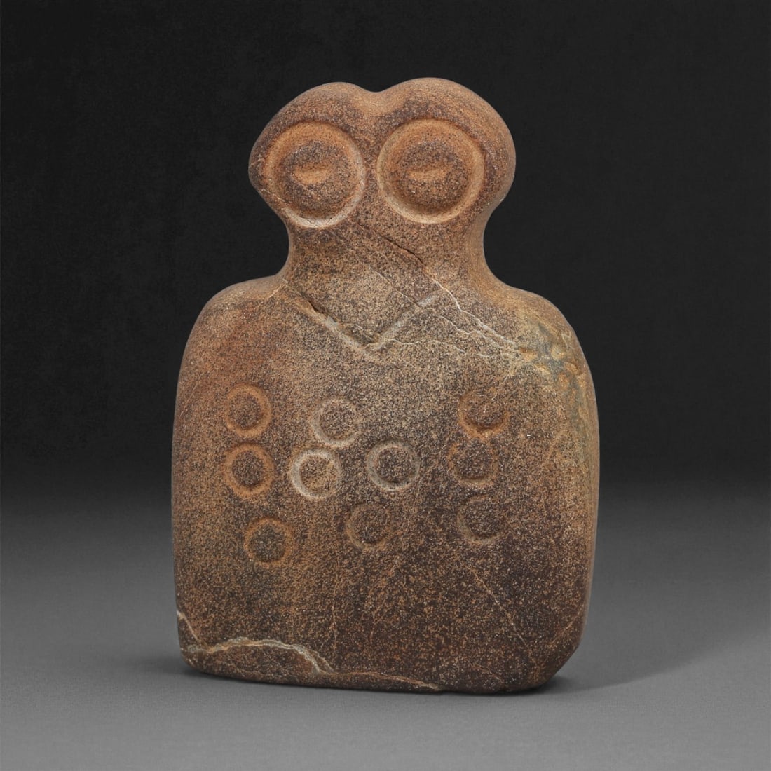 A Near Eastern Stone Eye Idol, 4th-3rd Millennium BCE, 32ZA: Carved from a brown stone with lighter surface veining, the figure is shaped with a rounded upper section bearing two recessed circular eyes formed by concentric grooves. The body is rectangular in ou