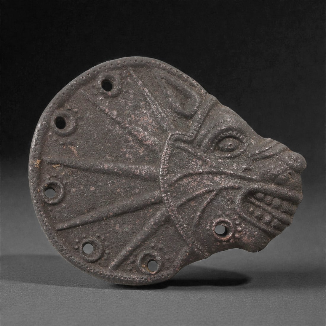 An Anatolian Iron Age Bronze Plaque with Lion Head, 8th-6th Century BCE, 61LP: Cast in bronze, the plaque is formed as a stylized lion head set against a flaring semicircular field. The face is defined by incised contour lines indicating the brow, cheeks, and an open mouth with 