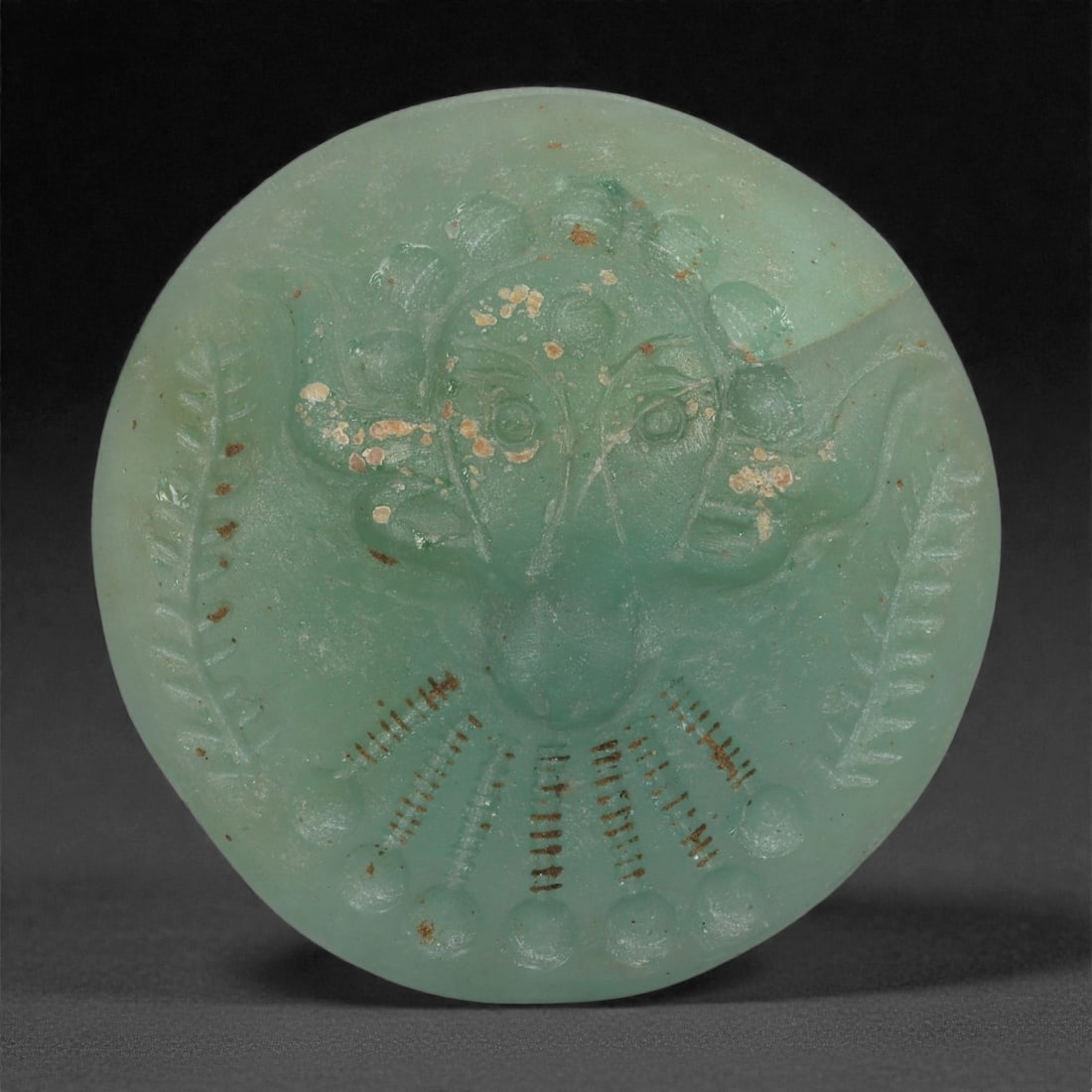 A Sasanian Green Glass Medallion with Anthropomorphic Motif, 3rd-7th Century CE, 27LP: Cast from translucent pale green glass, the medallion is circular in form and worked in shallow relief with an anthropomorphic mask featuring an elongated central snout and projecting lateral elements