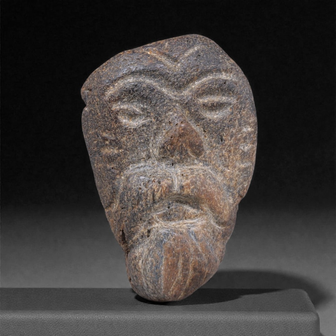 Ancient Fossil Bone Stone Anthropomorphic Bust - Miniature Head Sculpture, 5F: This small anthropomorphic bust is carved from dense fossilized bone stone and presents a solemn human face rendered in a strongly archaic style. The elongated head, heavy brow, deep-set eyes, and pro