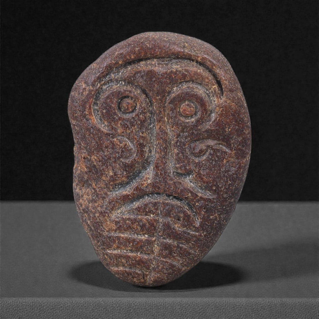 Ancient Fossil Bone Stone Anthropomorphic Maskette - Miniature Face Effigy, 28F: This miniature anthropomorphic maskette is carved from dense fossilized bone stone and presents a highly stylized human face rendered in a compact, oval format. Despite its small scale, the carving co