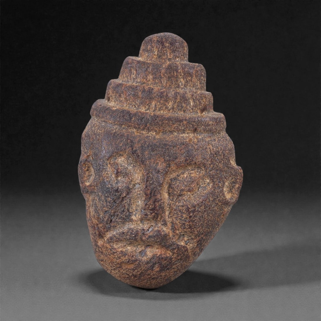 Ancient Fossil Bone Stone Anthropomorphic Head Effigy - Tiered Headdress Sculpture, 92E: This compact anthropomorphic effigy represents a human head crowned with a stepped, tiered headdress, carved from dense fossilized bone stone. The face is rendered in a highly stylized, archaic manner