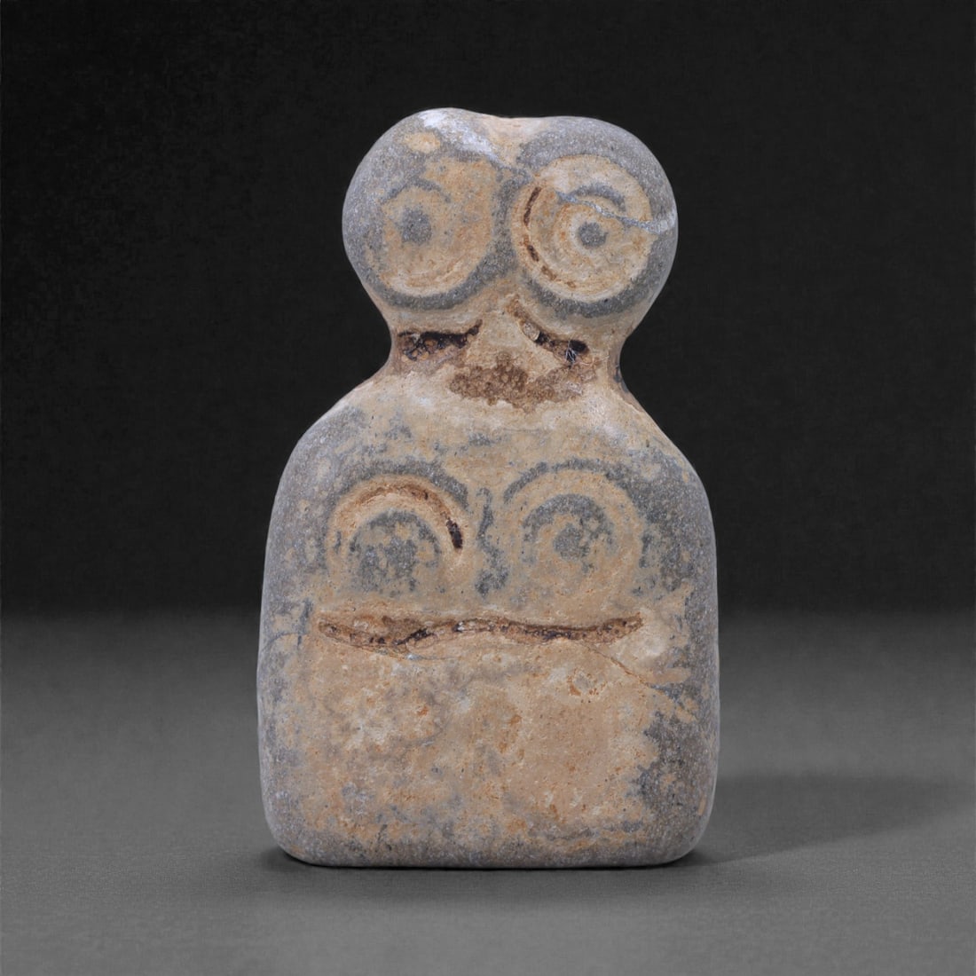 A Near Eastern Steatite Eye Idol, 4th-3rd Millennium BCE, 69L: Carved from grey-beige steatite, the figure is shaped with a rectangular lower body and a rounded upper section bearing a pair of prominent circular eyes. Additional concentric incised rings emphasize