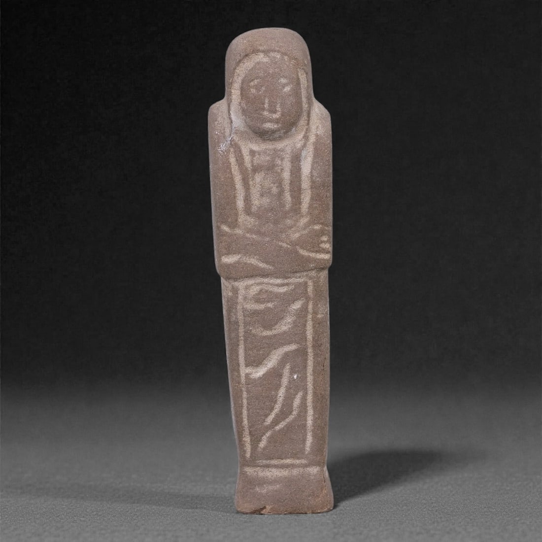 Egyptian Revival Stone Idol - Mummiform Figure, 68F: Carved from a compact brown stone, this mummiform figure is rendered in a frontal, elongated form with a rounded head, softly modeled facial features, and schematically indicated arms folded across th