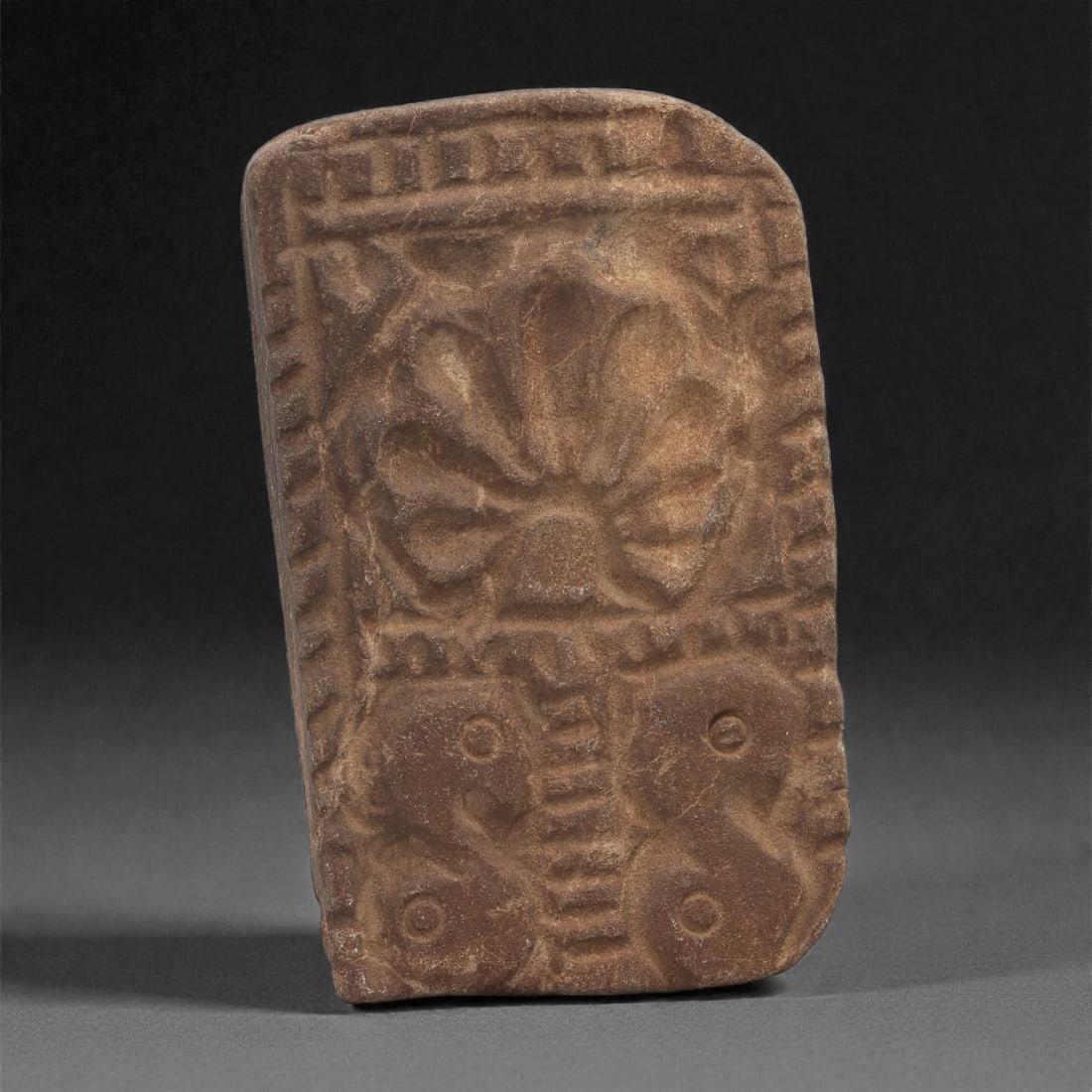 A Byzantine Stone Relief Plaque with Floral and Serpentine Motifs, 11th-12th Century, 26B: Carved from a reddish-brown stone with lighter surface veining, the plaque is organized into two stacked registers framed by a shallow recessed border. The upper register contains a stylized rosette w