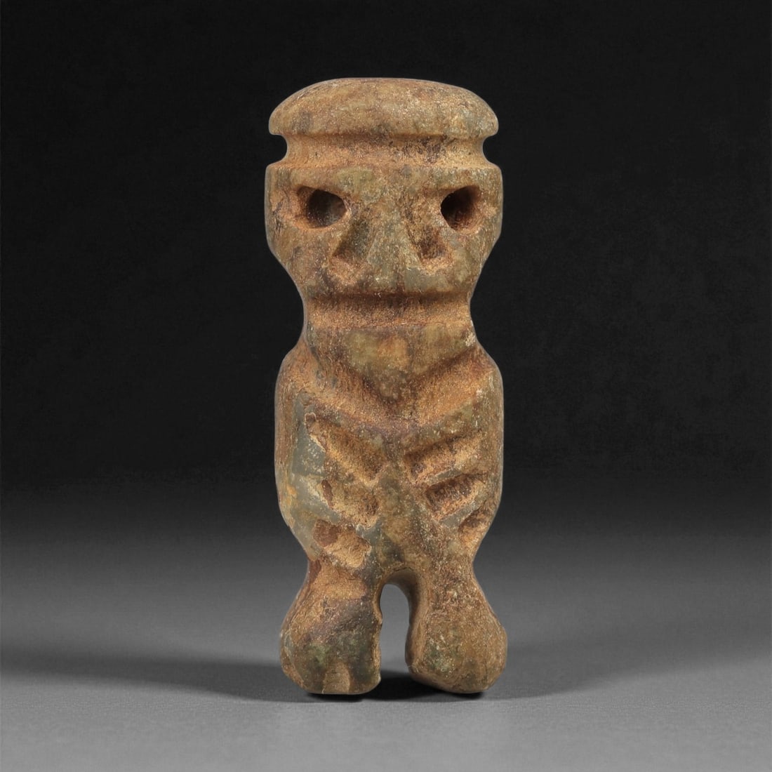 A Taino Carved Stone Idol, Late Ceramic Age, circa 1200-1500 CE, 69LE: Carved from mottled brown stone with areas of greenish accretion, the figure is modeled standing with short legs separated by a narrow gap. The head is rendered with a flat upper surface and recessed