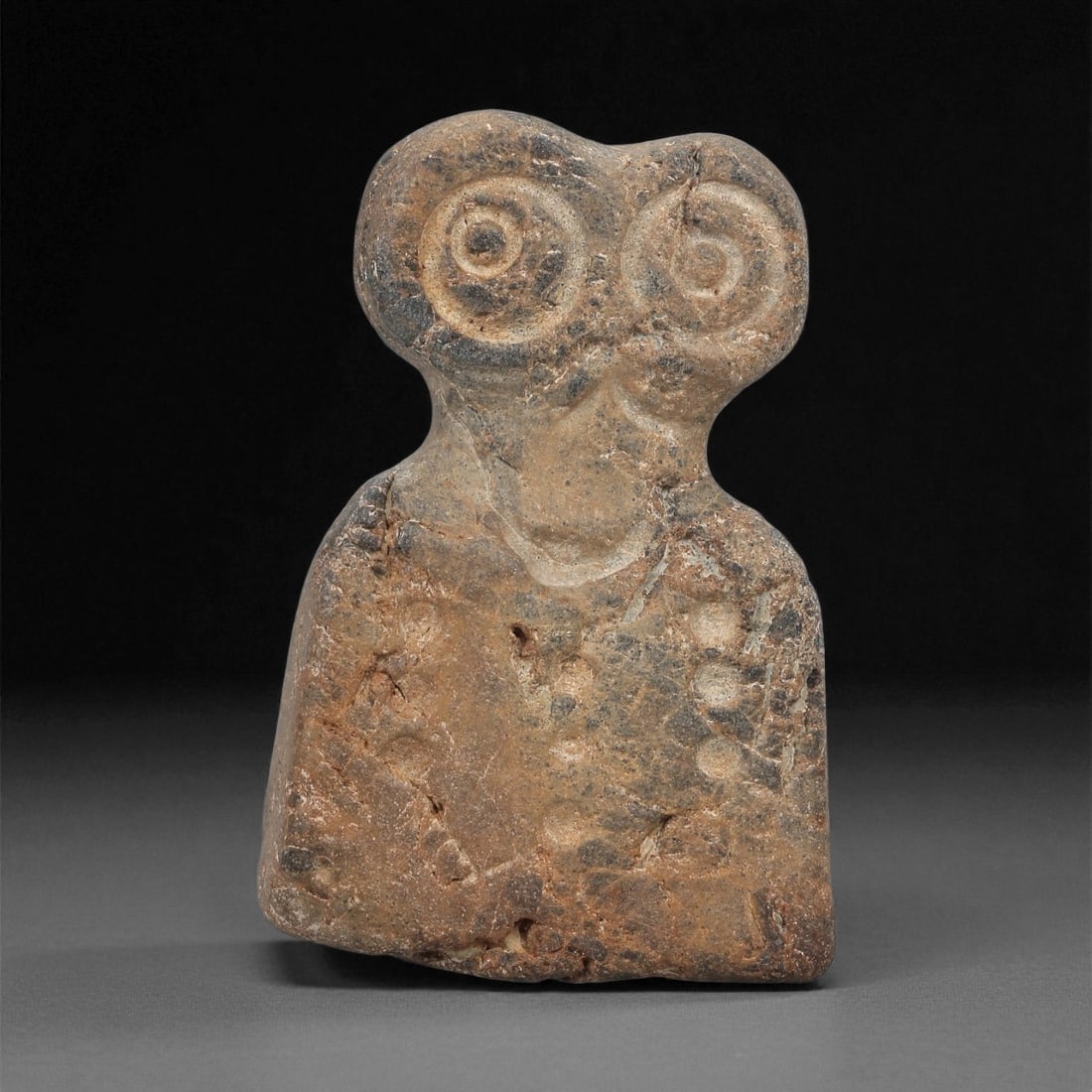 A Near Eastern Stone Eye Idol, 4th-3rd Millennium BCE, 56LB: Carved from a brown stone with darker surface inclusions, the object has a tapering rectangular body supporting a narrow neck and a bifurcated upper section. The head bears a pair of recessed circular