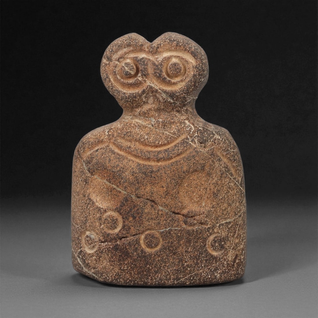 A Near Eastern Stone Eye Idol, 4th-3rd Millennium BCE, 60LB: Carved from a brown stone with lighter surface veining, the object is formed with a rounded rectangular body, a narrow neck, and a bifurcated upper section. The head bears a pair of recessed circular