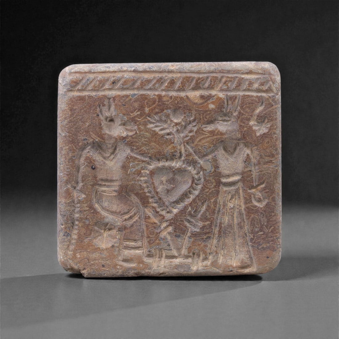 An Akkadian Fossilized Stone Stamp Seal, Circa 24th-22nd Century BCE, 77L: Carved from fossilized stone with variegated brown and grey tones, the seal is square in plan with a flat engraved face and a slightly rounded upper surface. The engraved field depicts two standing hu