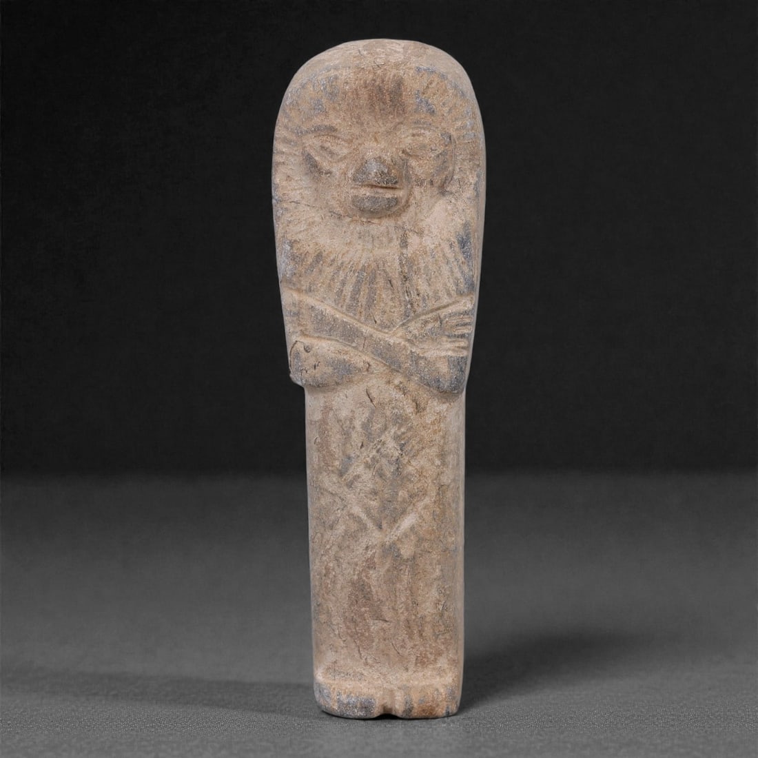 Egyptian Revival Stone Idol - Mummiform Figure, 75F: Carved from a mottled grey-brown stone, this mummiform figure is presented in a frontal, elongated form with a rounded head, stylized facial features, and arms crossed over the chest in low relief, cl
