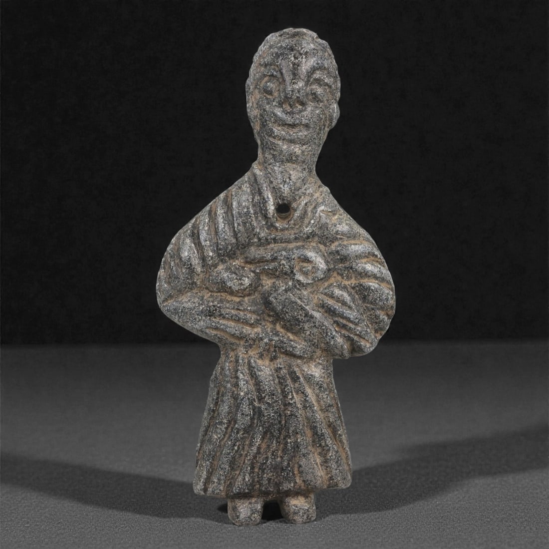 A Levantine Schist Anthropomorphic Amulet, Iron Age II, 9th-7th Century BCE, 100LJ: Carved from dark grey schist, the figure is shown standing frontally with short legs, a rounded head, and incised facial features including recessed oval eyes and a pronounced mouth. The body is envel