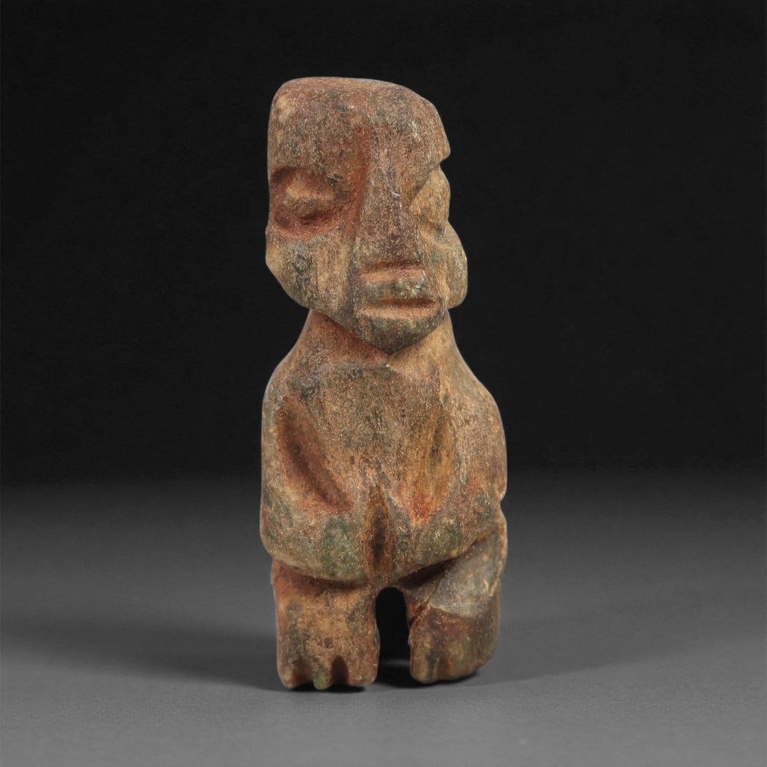 A Taino Carved Stone Idol, Late Ceramic Age, circa 1200-1500 CE, 23LD: Carved from mottled brown-green stone with surface weathering, the figure is shown standing with short bifurcated legs and a compact, tapering body. The head is rectangular with recessed eyes, a sharp