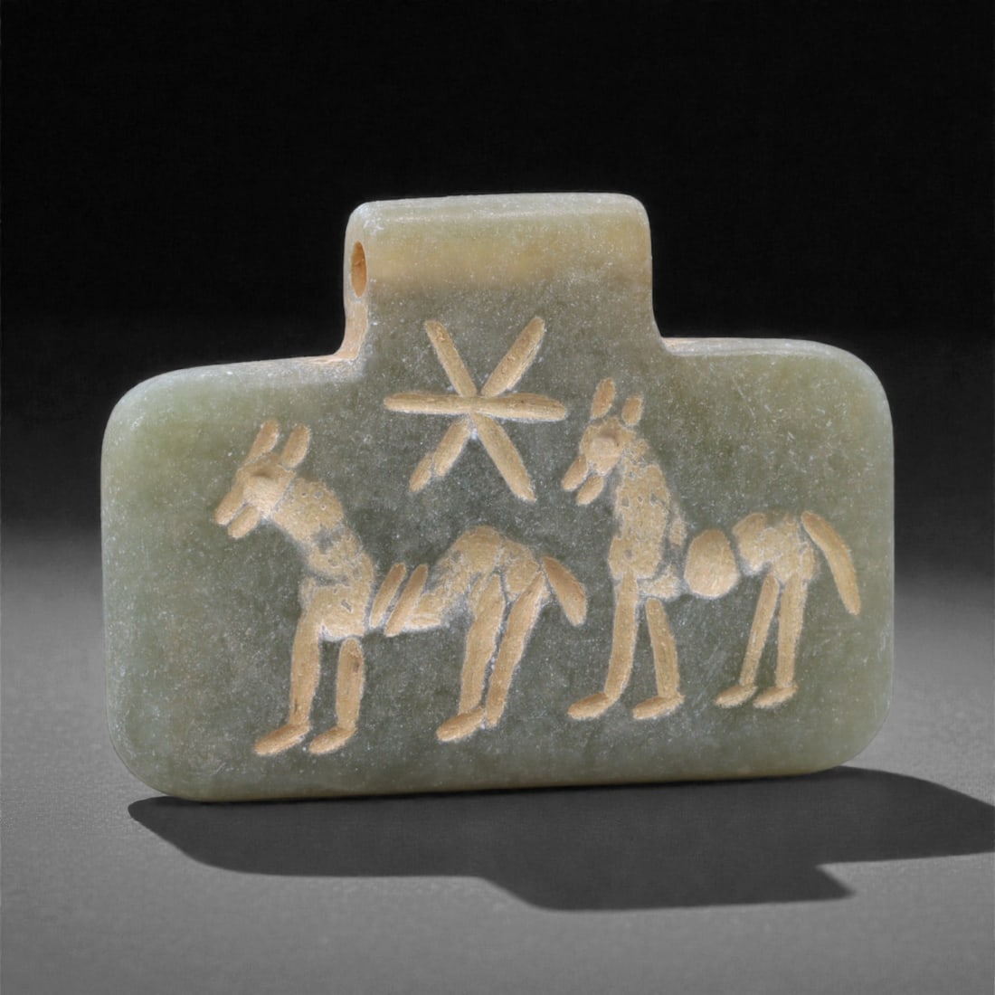 An Eastern Iranian Serpentine Amulet, Islamic Period, 9th-10th Century CE, 3.4cm, 68Q: Carved from pale green serpentine with a smooth, slightly translucent surface, the object is worked in low relief on one face, with the reverse left plain and undecorated. The carved elements are defi