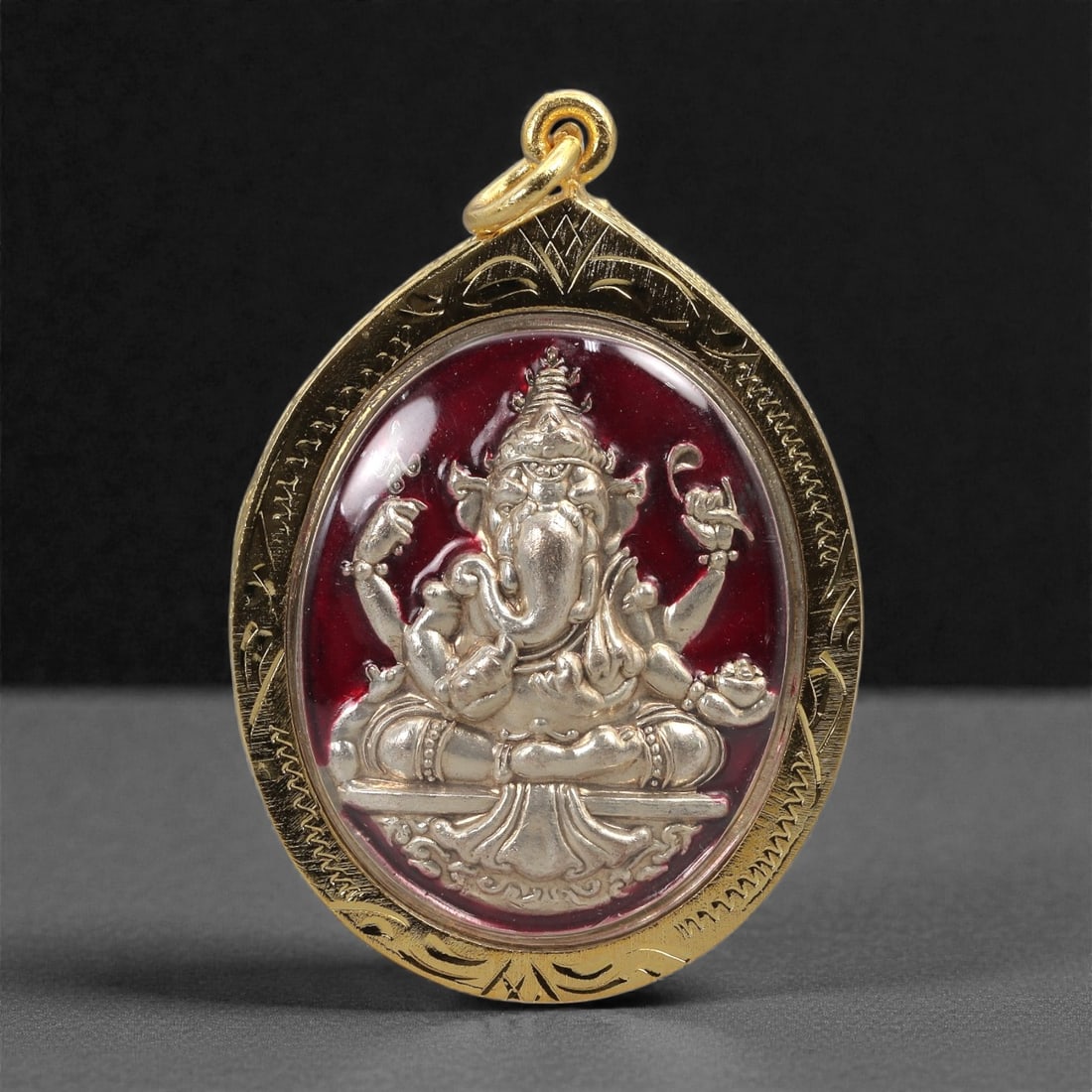 Hindu Metal Amulet - Ganesha Relief Pendant, 34D: This large oval pendant features a finely cast metal relief of Ganesha shown seated with multiple arms arranged symmetrically around the central body. The figure is set against a deep red ground that 
