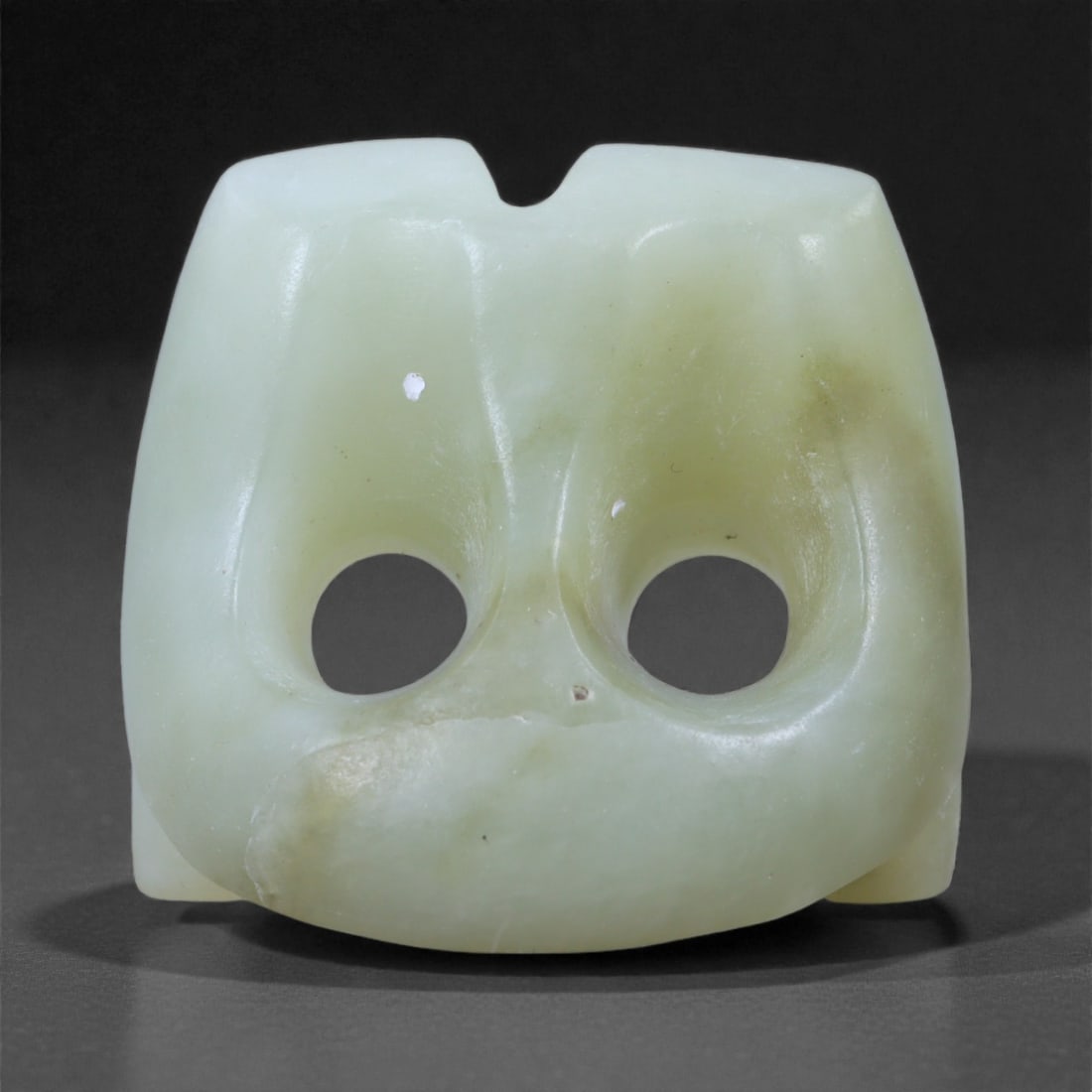 Chinese Jade Maskette, 19th-20th Century CE, 60L: Carved from pale green jade with minor cloudy inclusions, the small maskette is shaped with a broad, rounded lower contour and a flattened upper edge featuring a central notch. The front displays two