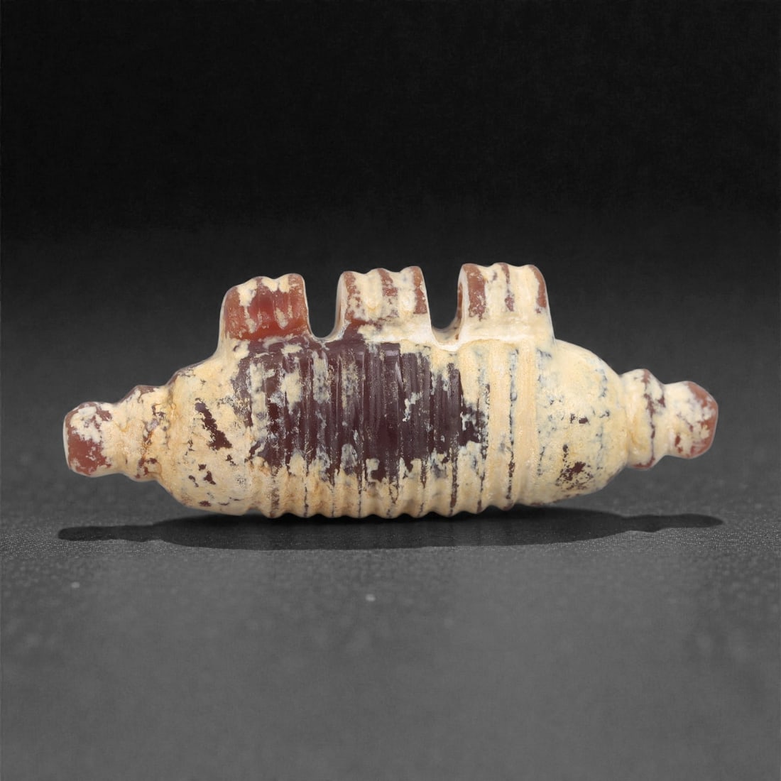 An Achaemenid Carnelian Amulet, 5th-4th Century BCE, 11A: Worked from mottled red carnelian with areas of pale surface accretion, the amulet is shaped as an elongated ribbed cylinder with regularly spaced transverse grooves along the body. Each end terminate