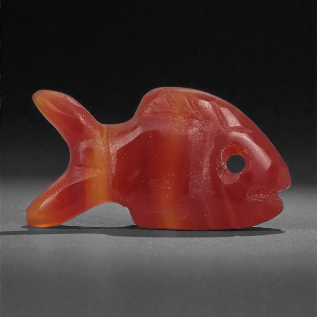 A Coptic Carnelian Fish Amulet, 4th-7th Century CE, 29LS: Carved from translucent red carnelian with warmer orange banding, the amulet takes the form of a stylized fish with a rounded head and a bifurcated tail. A circular perforation pierces the head for su