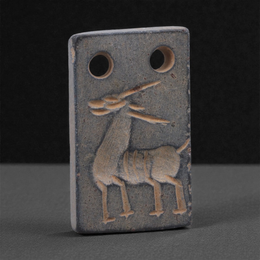 A Near Eastern Stone Double-Perforated Amulet, 3rd Millennium BCE, 94K: Carved from a fine-grained grey stone with minor surface wear, the amulet is formed as a small rectangular plaque with straight sides and slightly rounded edges. The upper edge is pierced with two cir