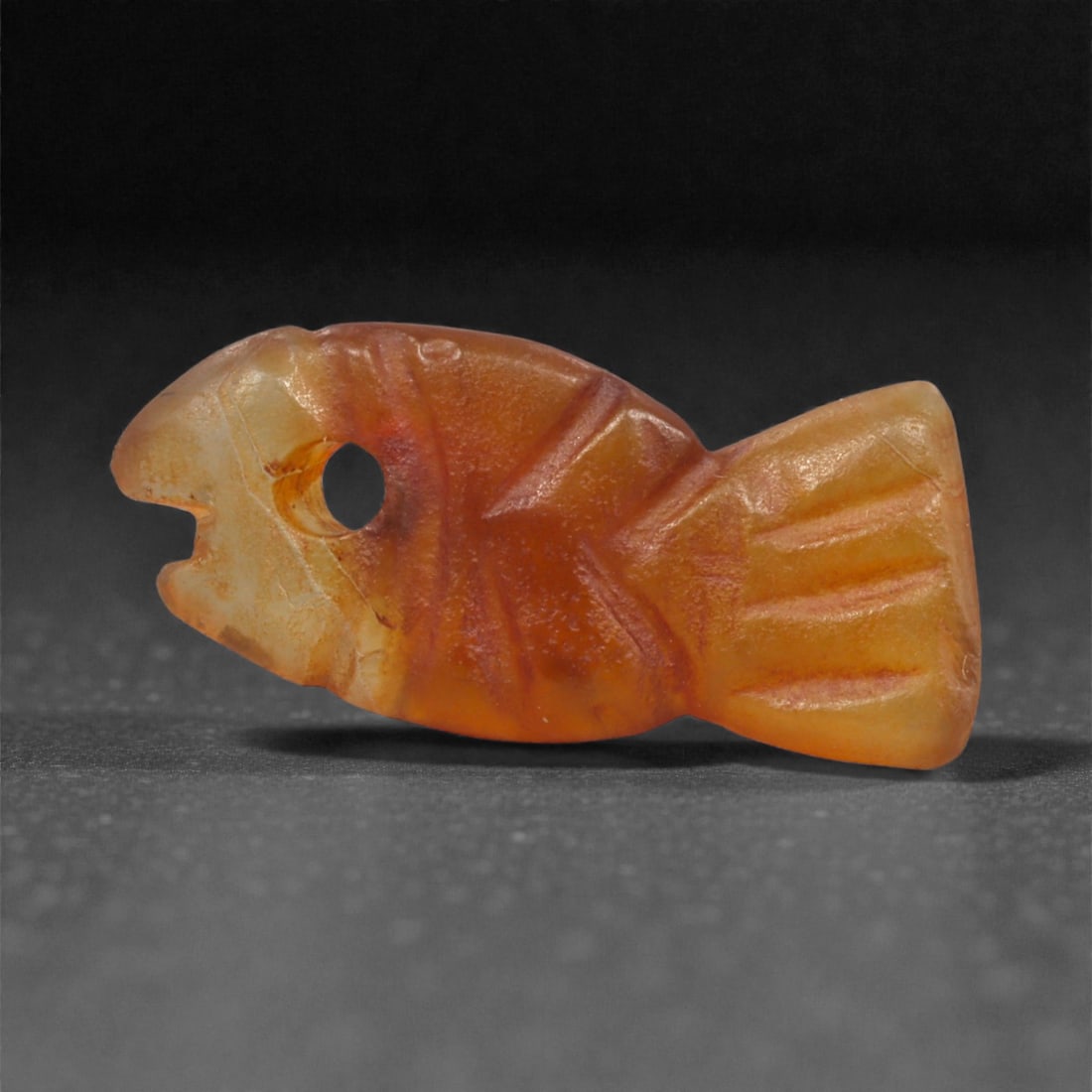 An Egyptian Carnelian Amulet, Coptic Period, 4th-7th Century CE, 2cm, 13LK: Worked from orange carnelian with natural internal variation, the amulet shows a compact form with smoothed contours and modest surface definition. Light wear is visible along the edges. Objects of th