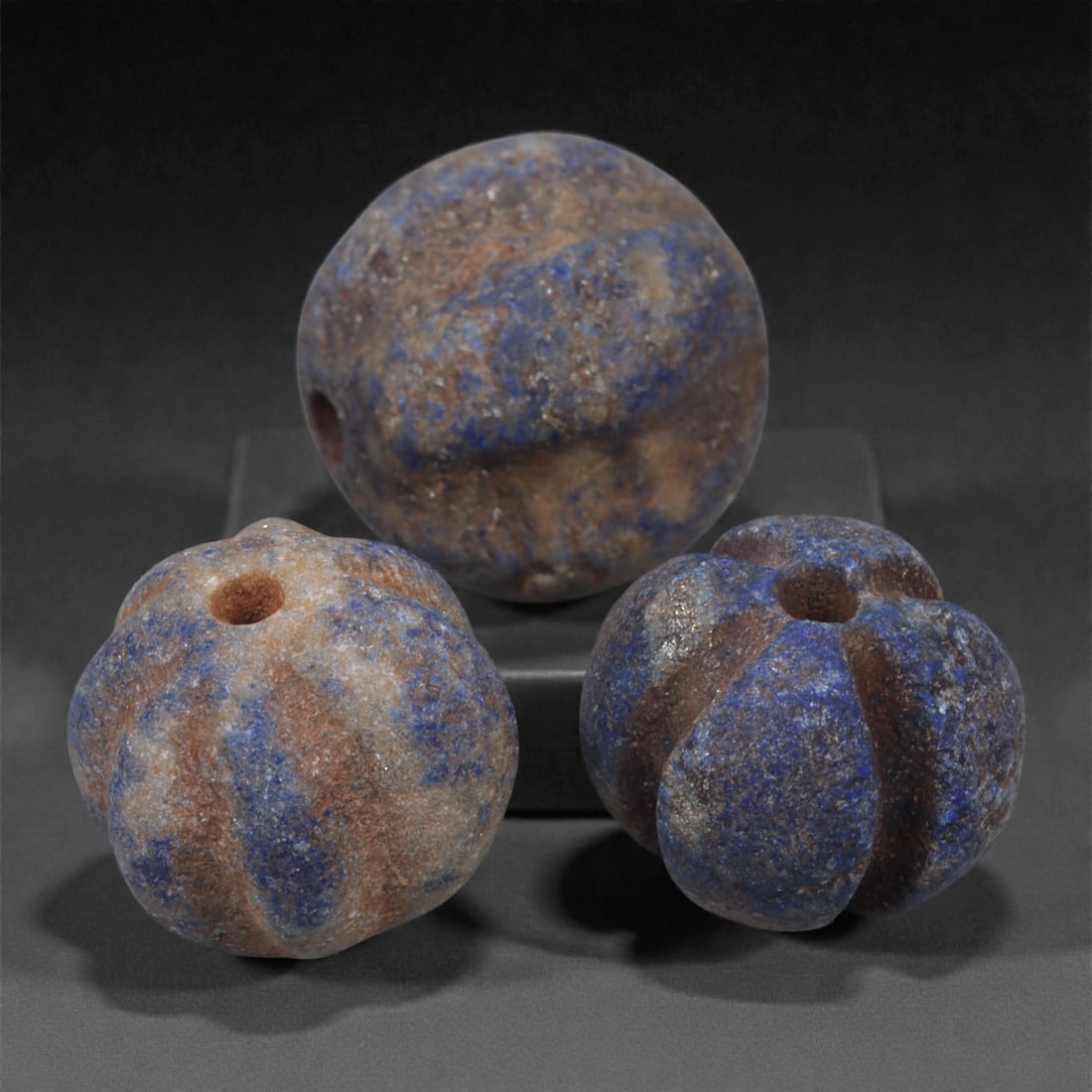 A Group of Three Egyptian Lapis Lazuli Melon Beads, 2nd Millennium BCE, 1.2cm, 11N: Carved from lapis lazuli with rich blue coloration interspersed with lighter calcite and brown mineral inclusions, the group comprises three melon-shaped beads of rounded, lobed form. Each bead is pie
