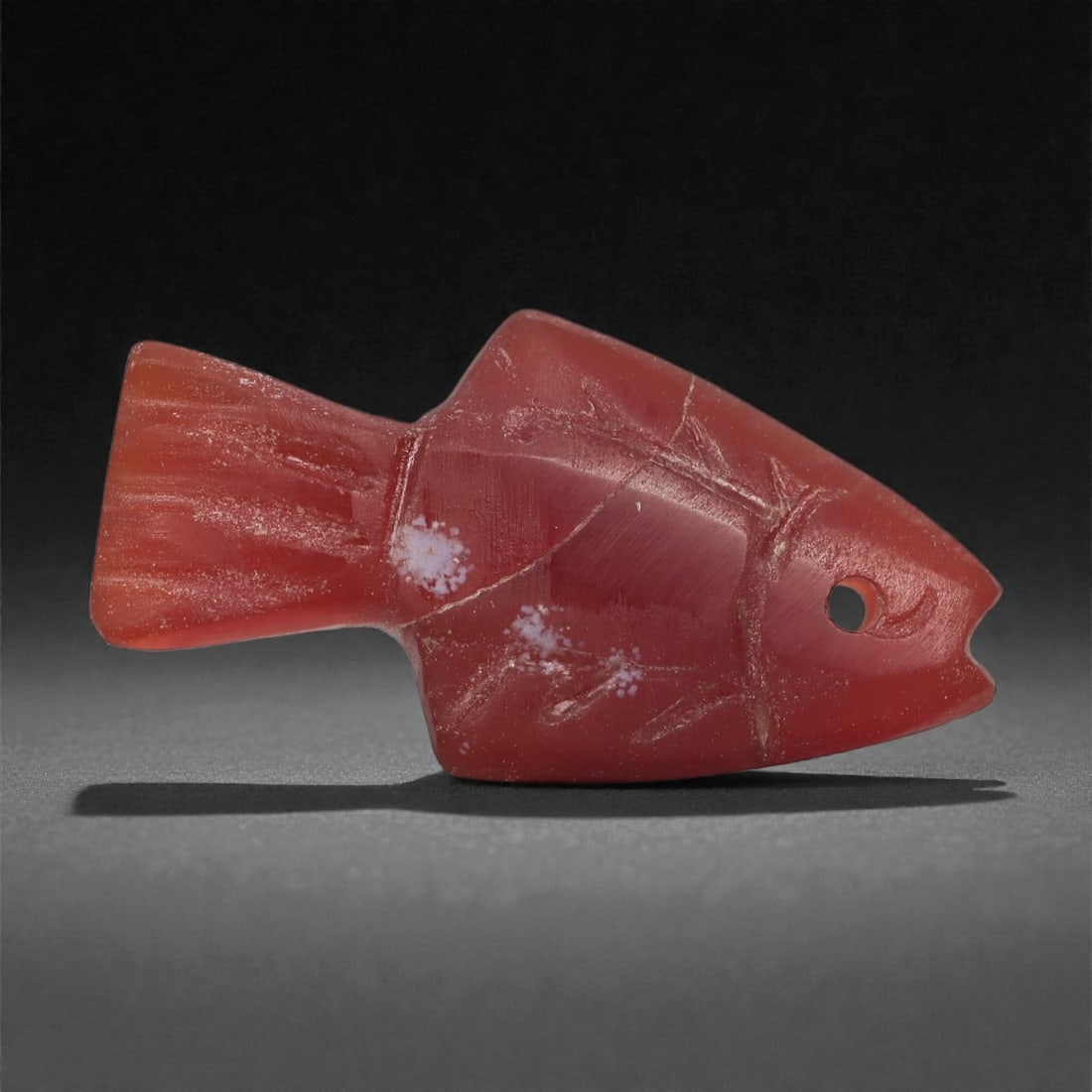 An Achaemenid Carnelian Fish Amulet, 5th-4th Century BCE, 24LS: Carved from translucent red carnelian with lighter internal veining, the amulet is shaped as a stylized fish with a squared tail, angular dorsal contour, and a notched mouth. A circular perforation at