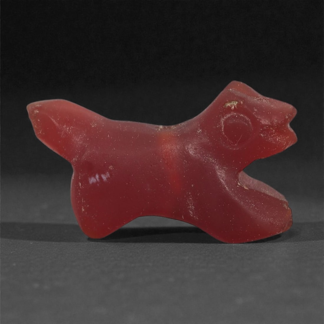 A Near Eastern Carnelian Canid Amulet, 1st Millennium BCE, 99LS: Carved from opaque red carnelian, the amulet depicts a small canid with a pointed snout, open mouth, and a circular recessed eye. The body is compact with a raised tail and short legs indicated by sim