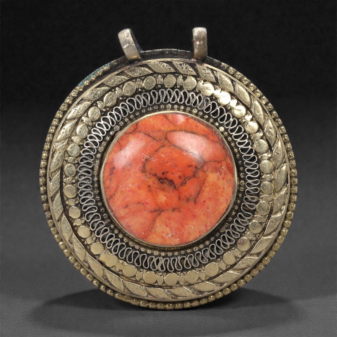 Himalayan Style Brass Pendant - Coral Cabochon Round, 9XF: This round brass pendant is constructed around a prominent coral cabochon whose mottled internal texture and warm orange-red coloration establish the visual focus of the piece. The stone is set within