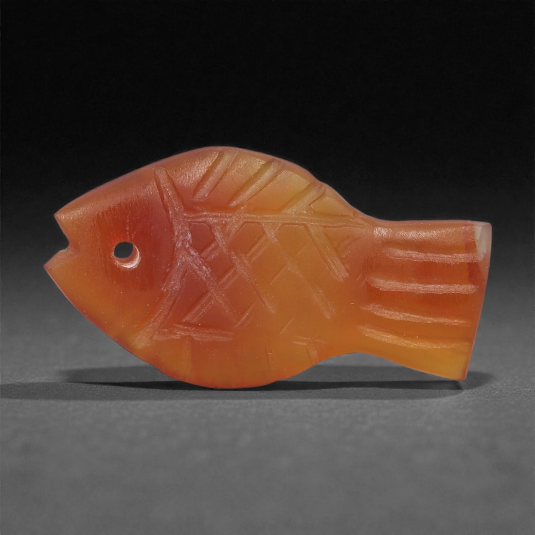 A Coptic Carnelian Fish Amulet, 4th-7th Century CE, 31LS: Carved from translucent orange carnelian with gradual tonal variation, the amulet is shaped as a stylized fish with a rounded body, squared tail, and a notched mouth. A circular perforation pierces th