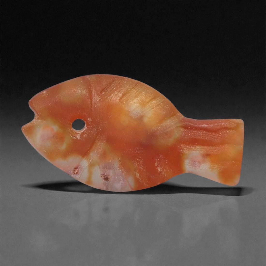 A Coptic Carnelian Fish Amulet, 4th-7th Century CE, 32LS: Carved from mottled orange carnelian with lighter cream and pink inclusions, the amulet is shaped as a stylized fish with an oval body, squared tail, and a notched mouth. A circular perforation at the