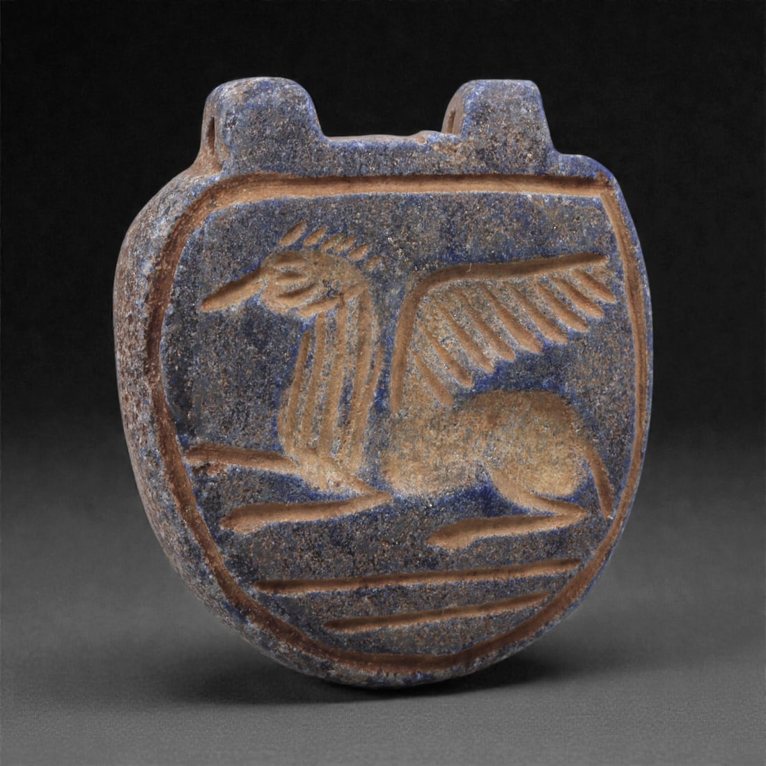 A Sasanian Lapis Lazuli Amulet, 3rd-7th Century CE, 3.3cm, 30K: Carved from deep blue lapis lazuli with visible lighter inclusions and natural granular texture, the object is finished with a flat reverse and a front surface worked in low relief. The carving is exe