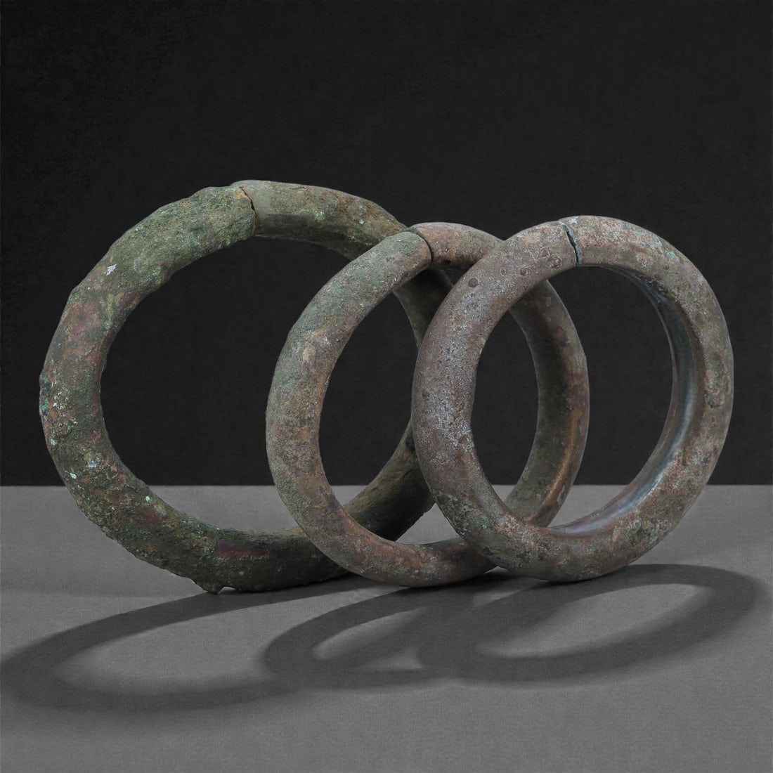 A Group of Roman Bronze Bracelets, 1st-3rd Century CE, 51H: Cast in bronze with circular-section profiles, the group comprises three open bracelets showing green and brown surface patination. Each is formed from a solid rod bent into an annular shape, the term