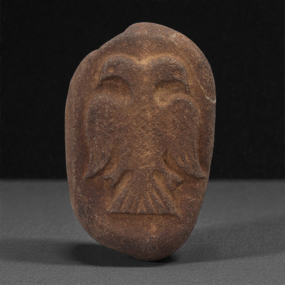 A Byzantine Stone Relief of a Double-Headed Eagle, 11th-13th Century, 68B: Carved from an ovoid pebble of reddish-brown stone, the relief depicts a double-headed eagle shown frontally with the two heads emerging from a shared neck. The wings are indicated by recessed fields