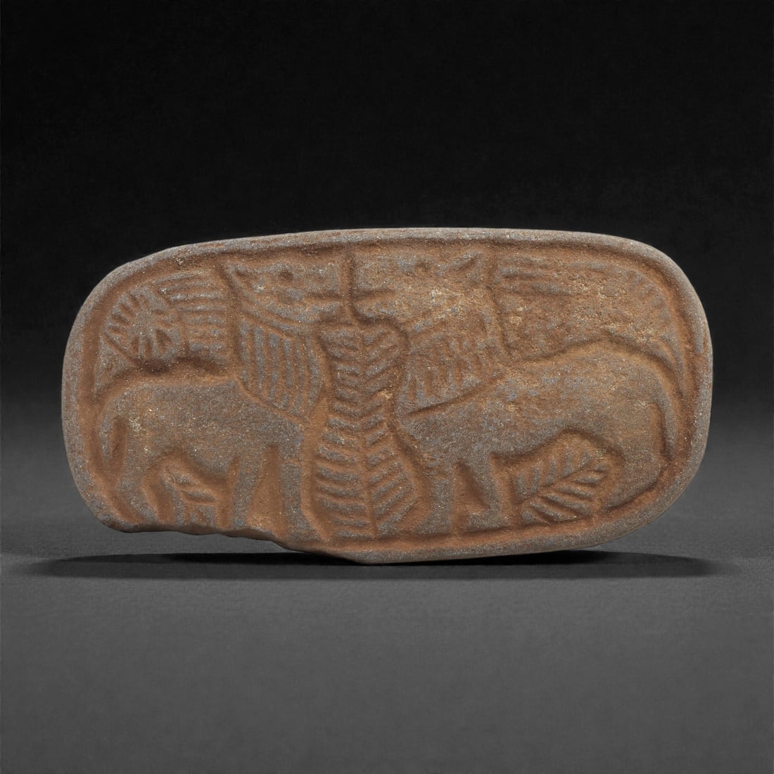 A Sasanian Lapis Lazuli Relief Plaque, 3rd-7th Century CE, 47C: Carved from a blue stone with brown surface accretions, the plaque is shaped as an elongated oval form with softly rounded edges. The front bears a shallow relief depicting two confronting animals arr