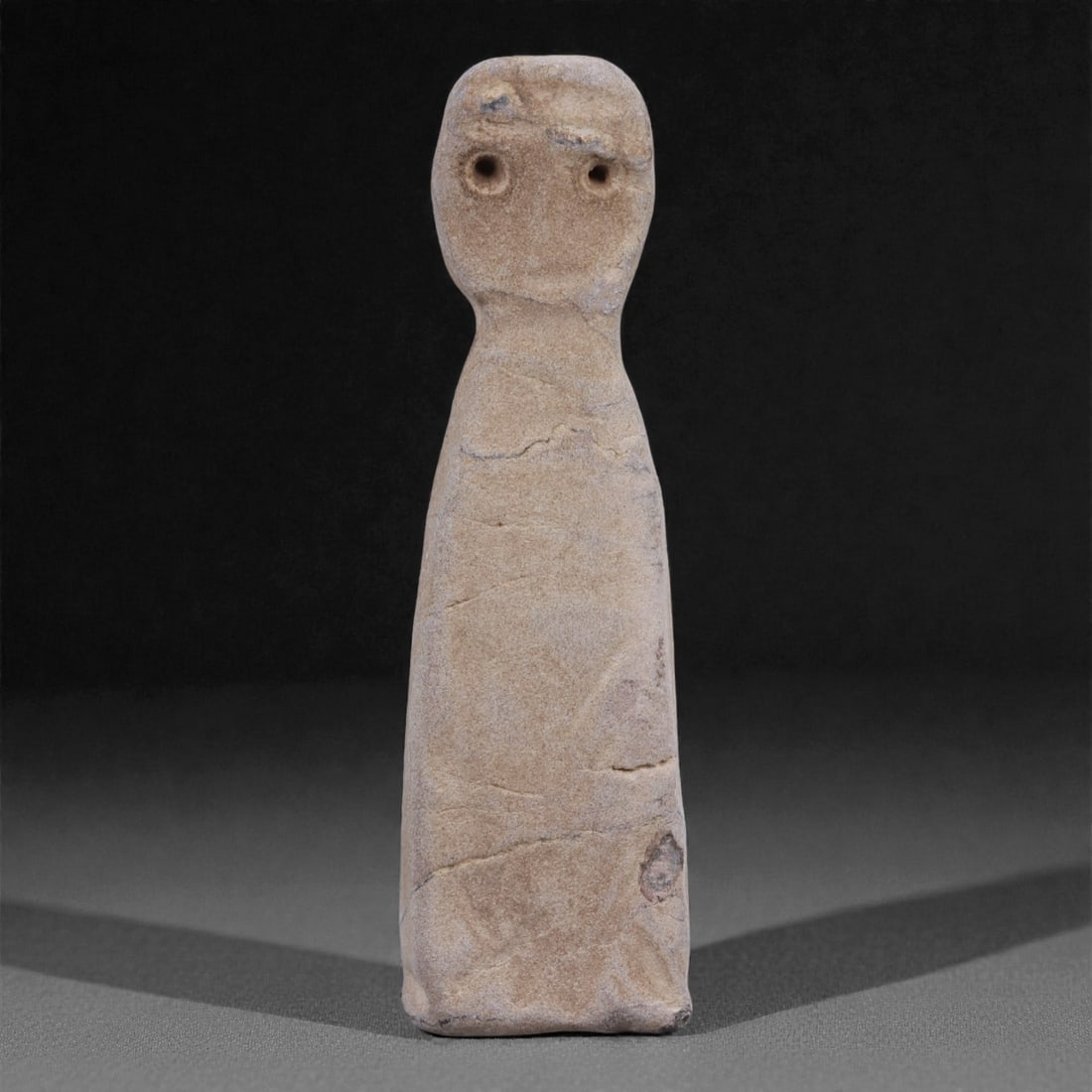 A Near Eastern Stone Anthropomorphic Figurine, 4th-3rd Millennium BCE, 99G: Carved from a light beige stone with fine granular texture and natural fissures, the object is formed as a standing anthropomorphic figurine with an elongated, columnar body and a flattened base. The