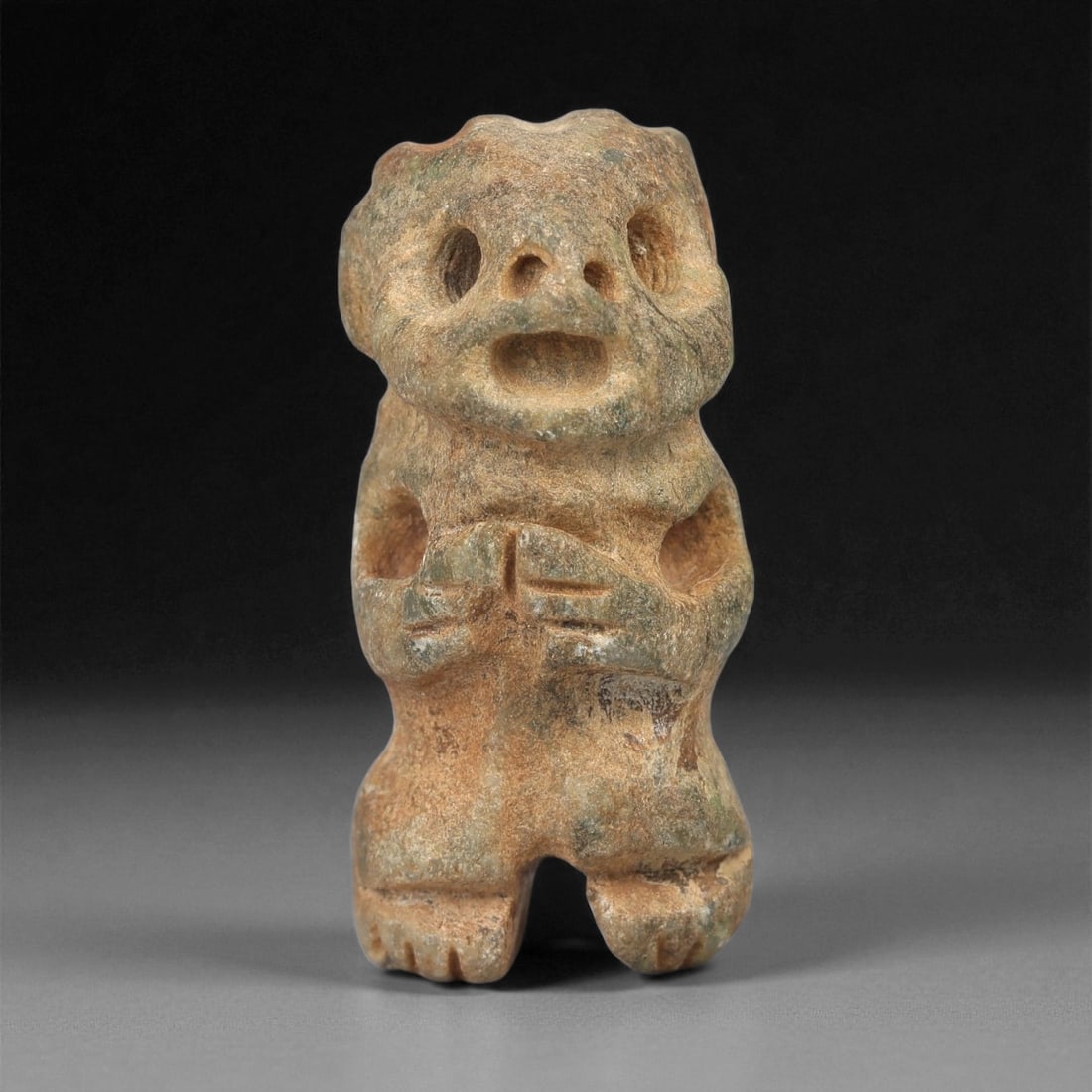 A Taino Carved Stone Idol, Late Ceramic Age, circa 1200-1500 CE, 64LE: Carved from mottled brown stone with traces of greenish surface accretion, the figure is modeled standing with short legs and a rounded torso. The head is broad, bearing recessed circular eyes above a